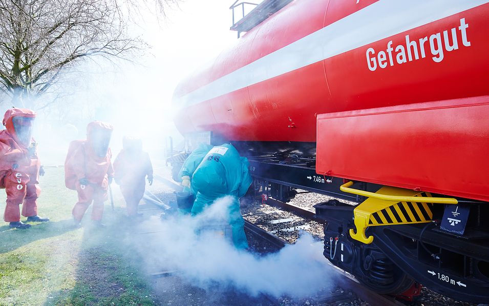 Fünf Mitarbeiter in Schutzkleidung trainieren vor rotem Kesselwagen.