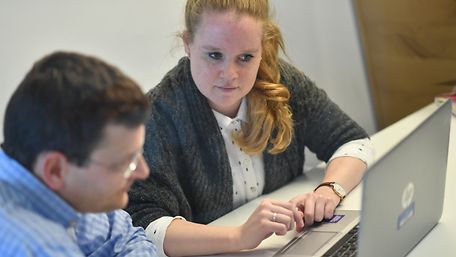 Junge Mitarbeiterin vor einem Laptop im Gespräch mit einem Kollegen