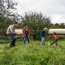 A family of four kick a ball around on a green field with a tank wagon in the background.